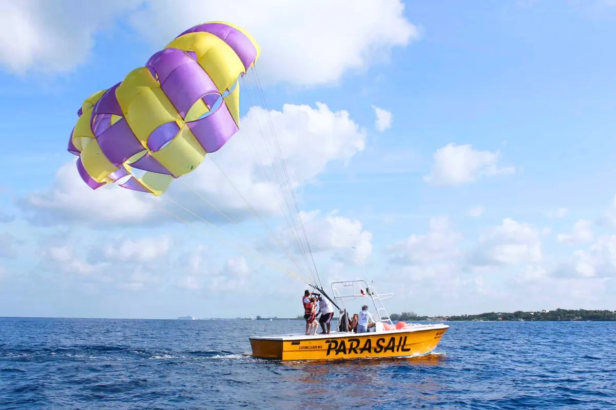 An orange boat marked "PARASAIL" on the side floats on the deep blue water off Cozumel, Mexico, with a large purple and yellow parachute rising into a partly cloudy sky as crew members on deck prepare a rider for takeoff.