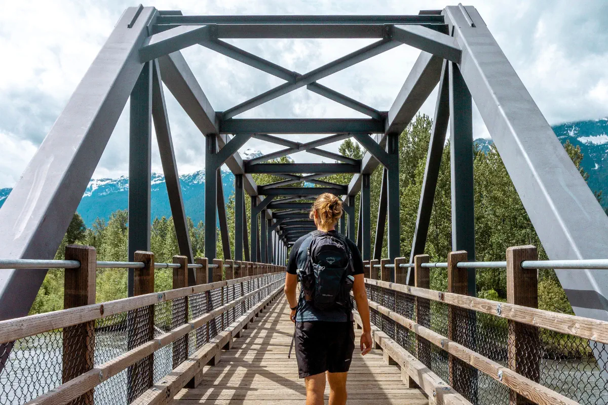 A person with a backpack walks across a steel and wood bridge surrounded by green trees, with snow-capped mountains visible in the background. The bridge’s symmetrical metal beams create a tunnel-like perspective leading toward the scenic nature in Revelstoke BC.