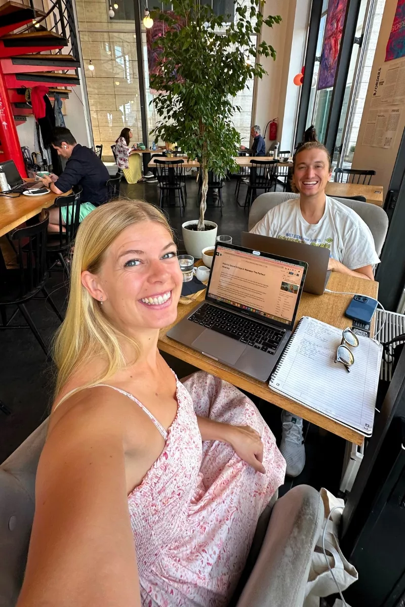 Two smiling people working on laptops in a modern, brightly lit café with high ceilings and large windows. The woman in the foreground is taking a selfie and has a notebook next to her. They both have coffees and laptops on the table.