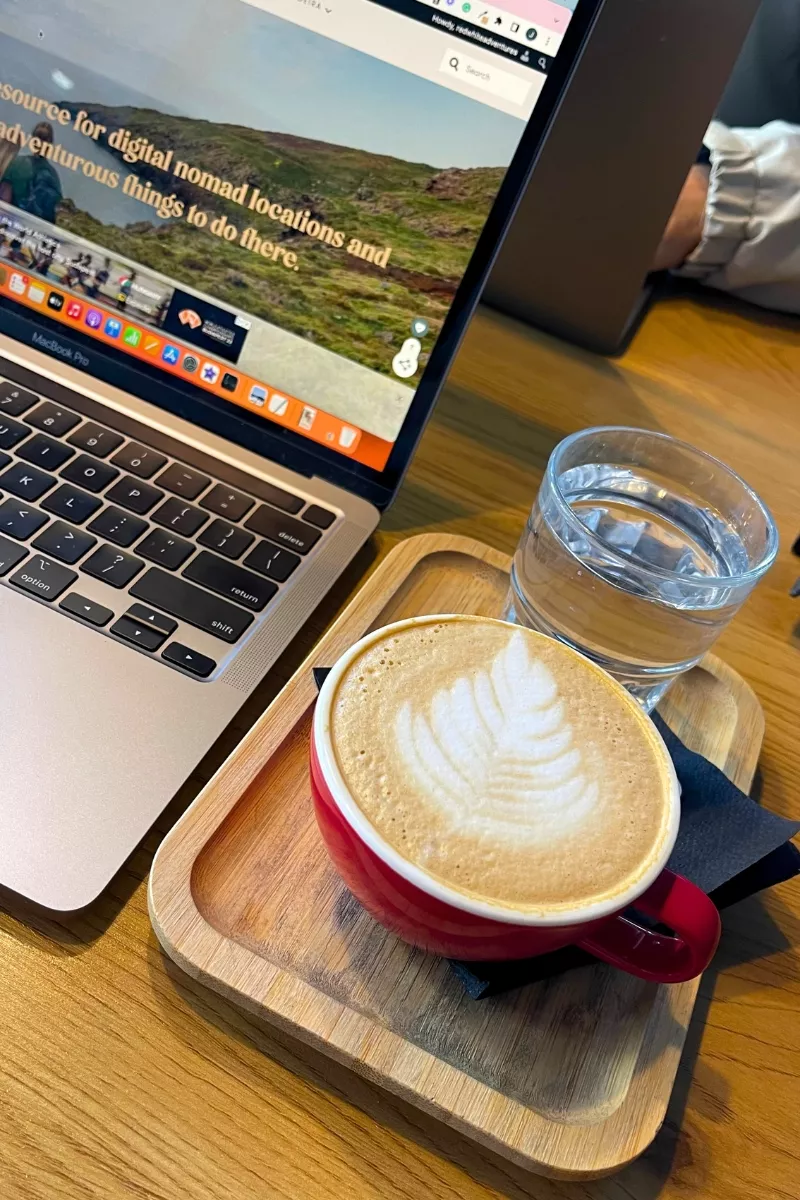 A close-up of a laptop on a wooden table beside a red cup of latte with leaf-shaped latte art and a glass of water on a small wooden tray. The laptop screen displays a scenic website about digital nomad locations.