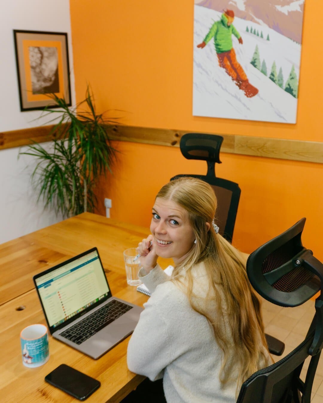 A woman smiles at the camera while working on a laptop at a wooden desk in a bright coworking space with orange walls. She sits in an office chair with a mug, phone, and notebook nearby, creating a relaxed digital nomad work setup.