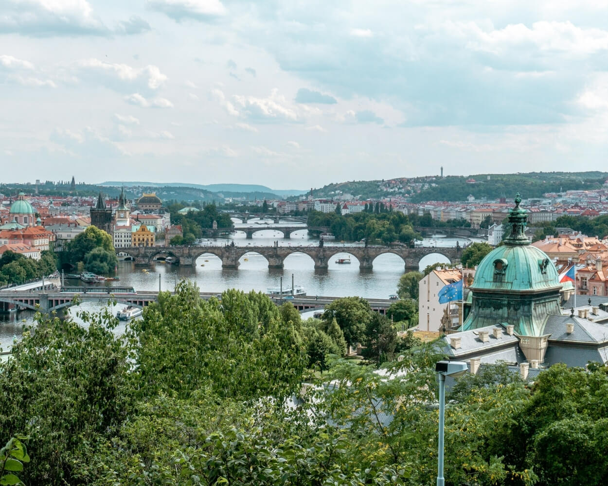 A panoramic view of Prague showcasing the Vltava River lined with historic stone bridges, including the iconic Charles Bridge. The city’s red rooftops, green copper domes, and tree-covered riverbanks frame the water.