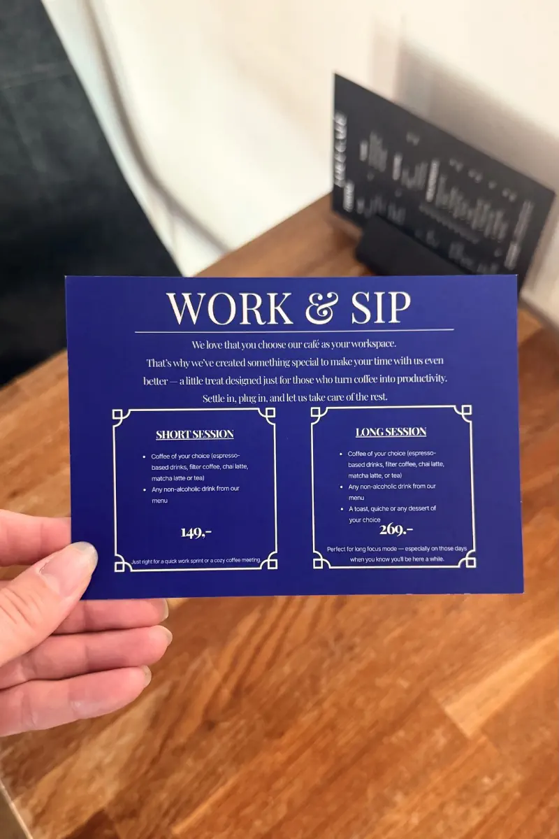 A hand holds a blue café card titled Work and Sip describing workspace packages on a wooden table. Text explains short session for 149 including a coffee and non alcoholic drink and long session for 269 including coffee a drink and a toast quiche or dessert.