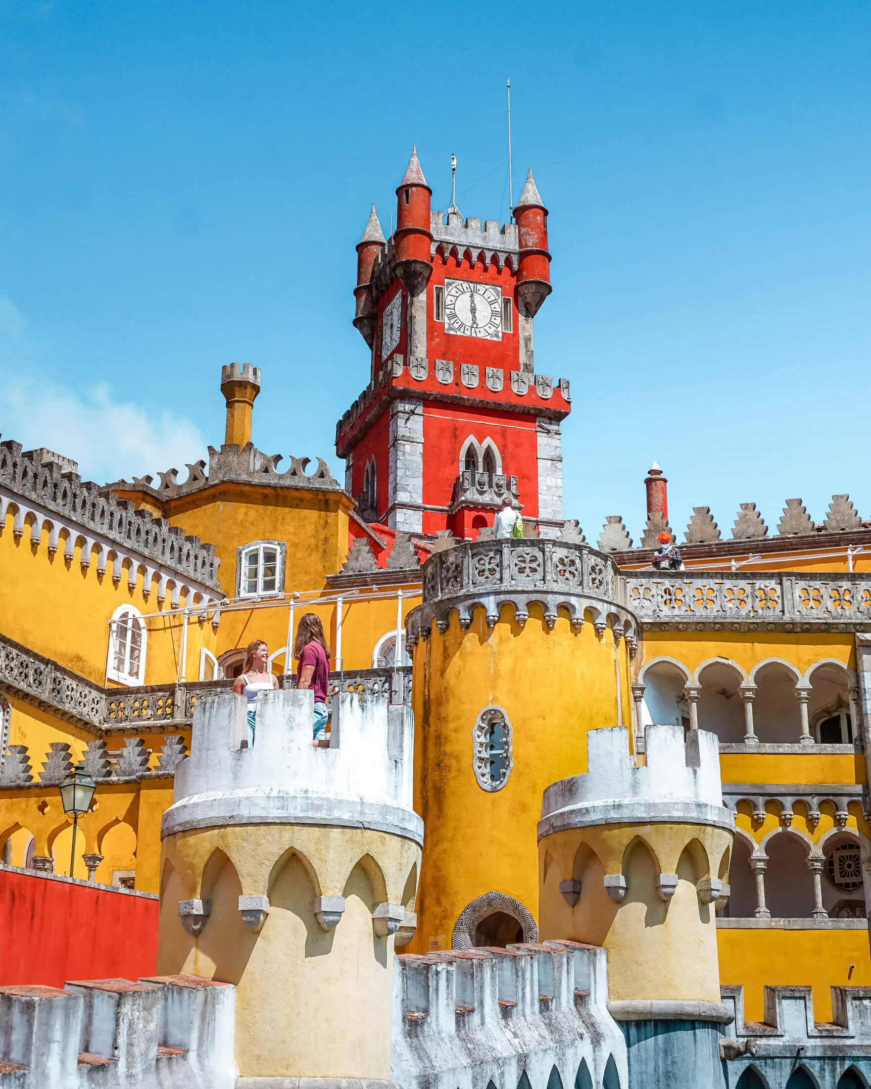 Colorful Pena Palace in Sintra with bright yellow walls and a red clock tower rising above decorative turrets and arches. Two visitors stand on a balcony overlooking the ornate architecture against a clear blue sky.