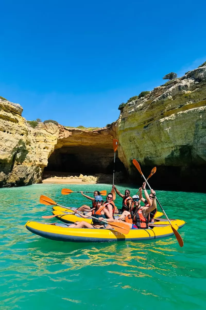 A group of people in bright yellow kayaks wearing life jackets and holding paddles, smiling in front of a sea cave with golden limestone cliffs and turquoise water. This Benagil cave kayak tour showcases the dramatic entrance of the famous cave under a clear blue sky.