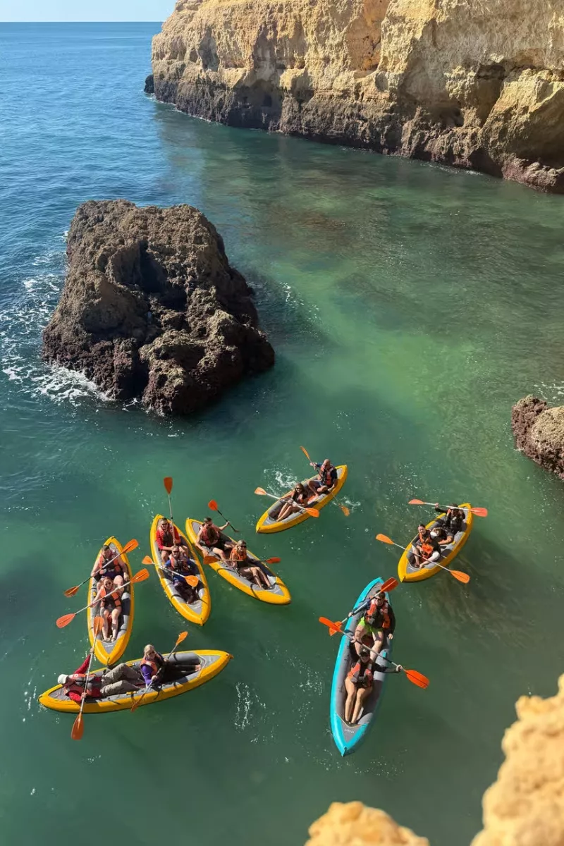 An overhead view of several kayaks with small groups of paddlers exploring calm green-blue water beside towering cliffs and sea stacks. The scene captures a kayak adventure along the Algarve coast.