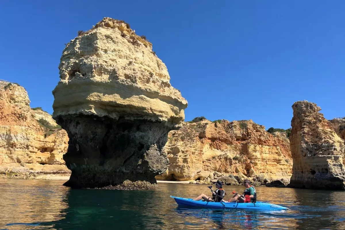 Two people paddle a bright blue kayak past a towering limestone rock formation in the sea, set against rugged Algarve cliffs and a clear blue sky. 