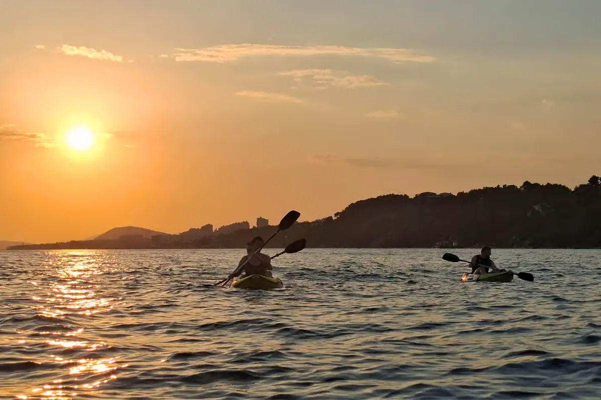 Two people in paddling kayaks on the open water off Split, Croatia, silhouetted against a vivid orange sunset with a hilly coastline stretching across the horizon behind them.