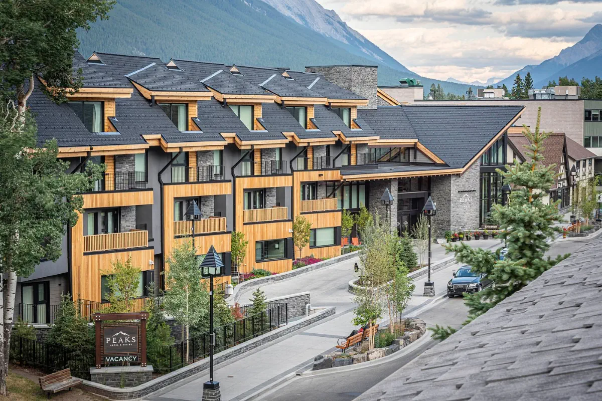 Exterior view of Peaks Hotel & Suites in Banff, Canada, featuring modern alpine-style architecture with wood and stone accents. The multi-storey building has balconies, landscaped walkways, and a “Vacancy” sign near the entrance, set against a backdrop of forested mountains.