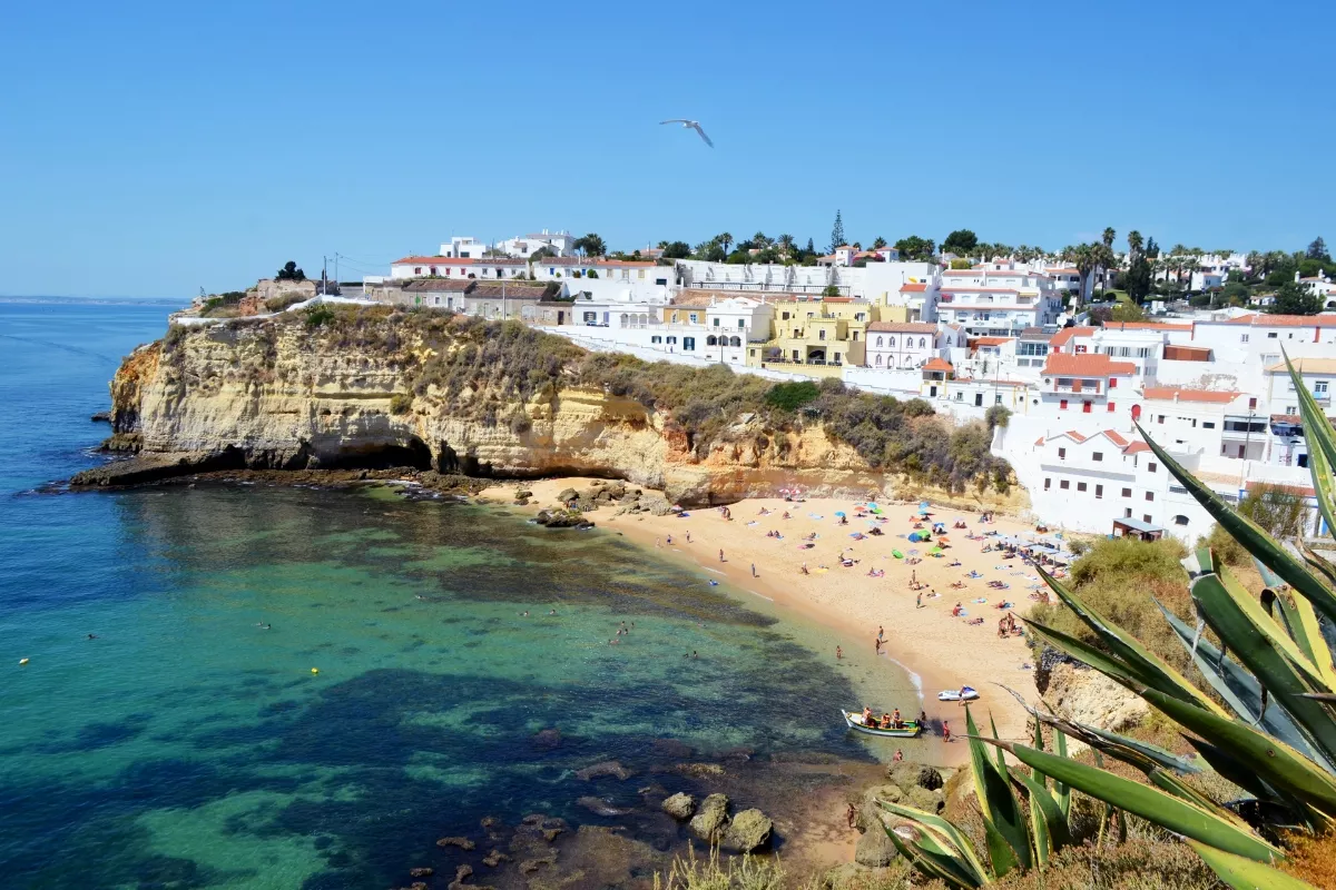Scenic coastal view of Carvoeiro in the Algarve (Portugal), featuring golden cliffs, turquoise waters, a sandy beach dotted with colourful umbrellas and whitewashed houses perched on the cliffs.