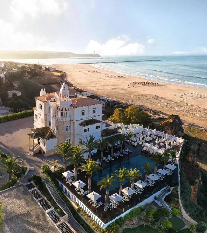 Aerial view of a grand beachfront hotel in Portimão with a central white villa, palm trees, and an outdoor pool and terrace overlooking a wide sandy beach and the ocean.
