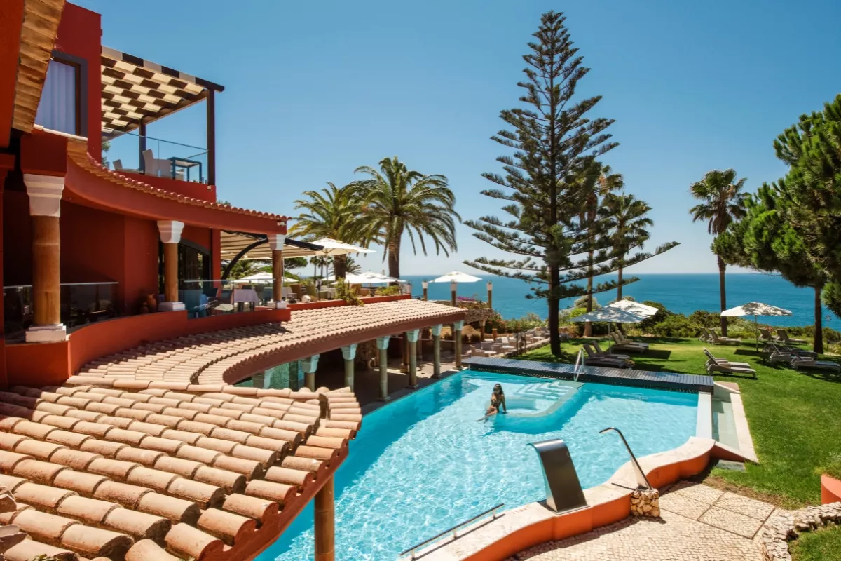 Outdoor swimming pool at a boutique hotel, complete with sun loungers, parasols, and ocean views framed by lush greenery and palm trees, and a view of the Atlantic Ocean.