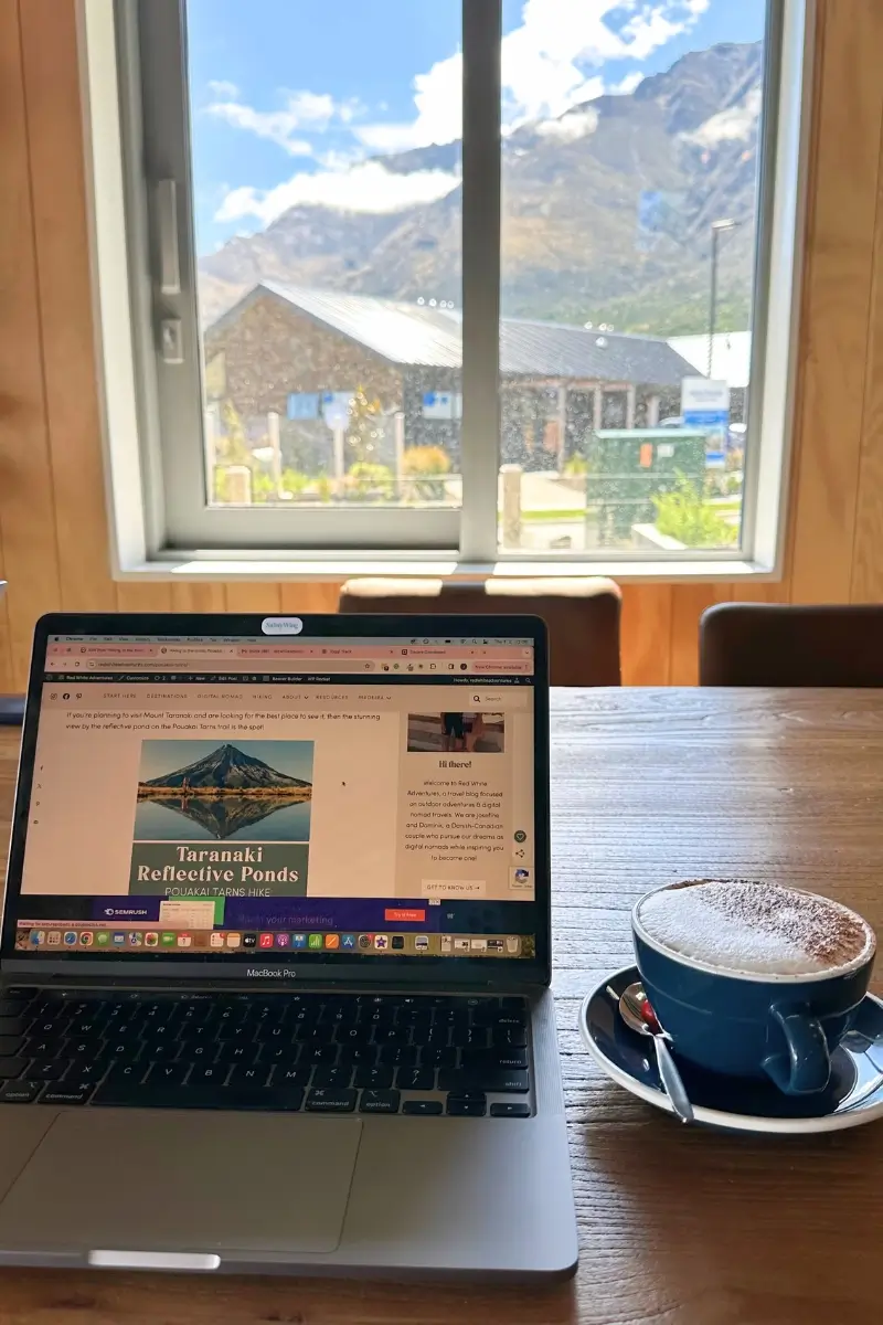 Blogging from a café in Queenstown, NZ. In the picture you see a laptop and a cup of coffee in front of a mountain view.