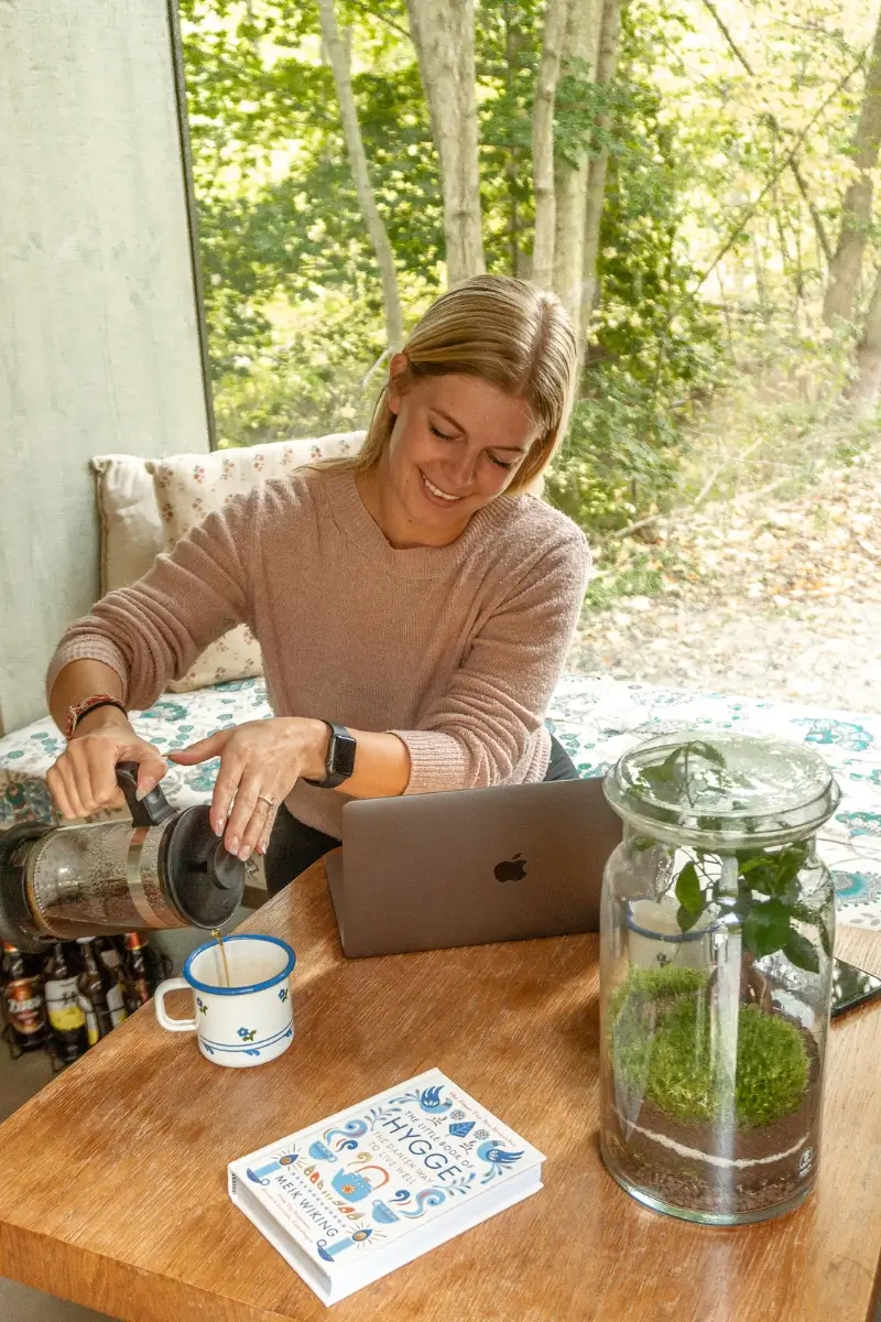 Young woman pouring herself coffee from a french press in front of her computer inside a cozy cabin with green trees in the background.