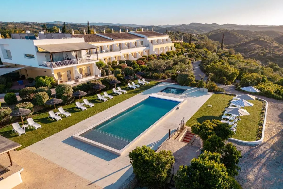 Aerial view of a rural hotel in the Algarve with a rectangular outdoor pool, surrounded by sun loungers and parasols, nestled in a lush green landscape with rolling hills in the background.