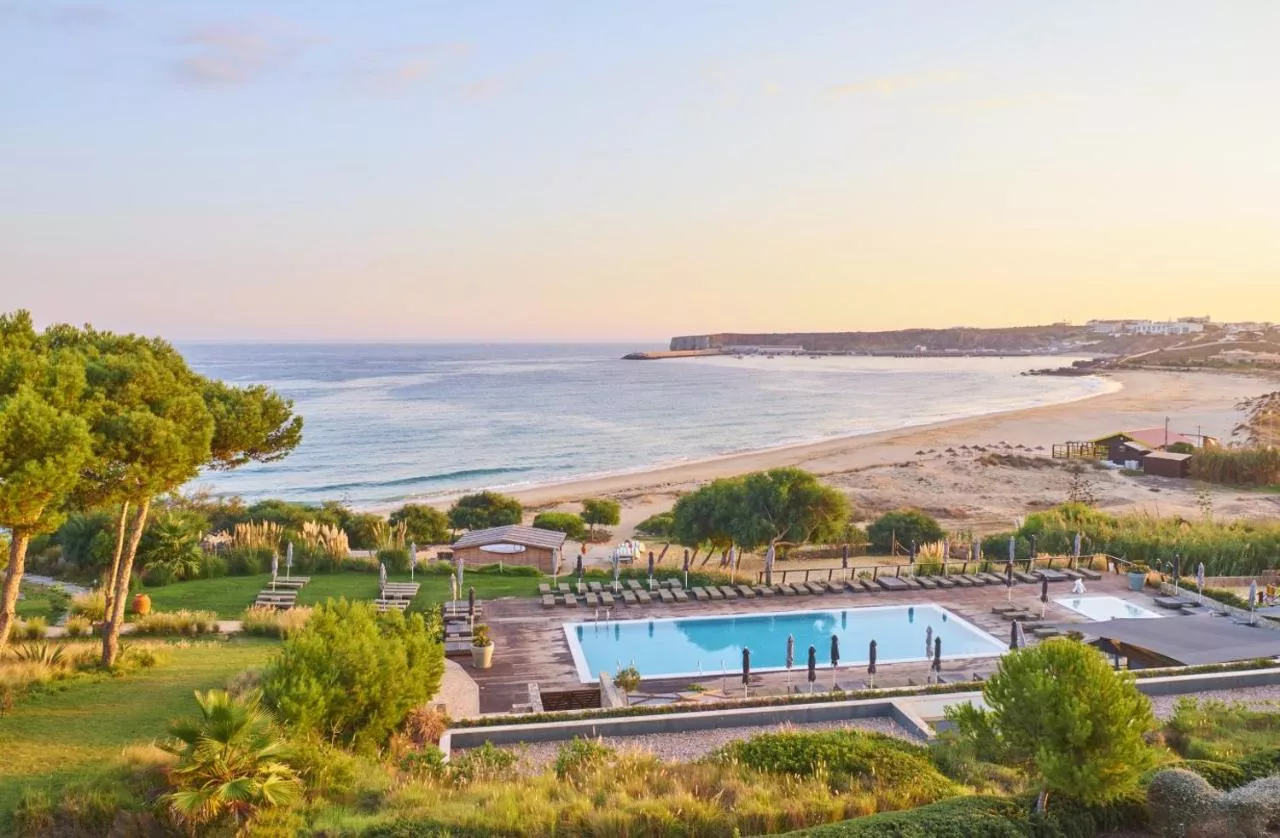 Aerial view of Martinhal Sagres Beach Resort in the Algarve, Portugal, with a pool area, sun loungers, lush gardens, and a sandy beach along the ocean at sunset.