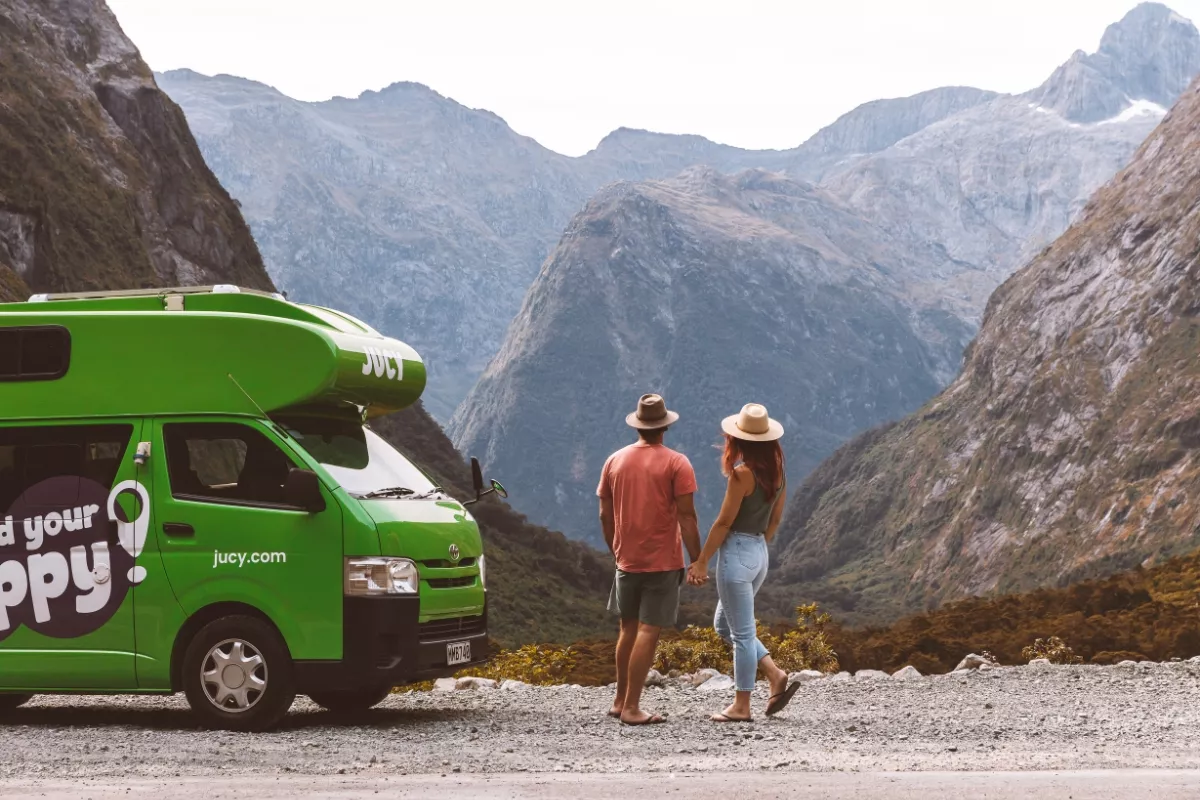 Lime-green JUCY campervan branded with “Find your happy!” and “jucy.com” is parked on a gravel pull-out along New Zealand’s Milford Road. A hat-wearing couple stand barefoot and hand-in-hand beside the van, admiring the sheer, slate-grey mountains that tower across the valley.