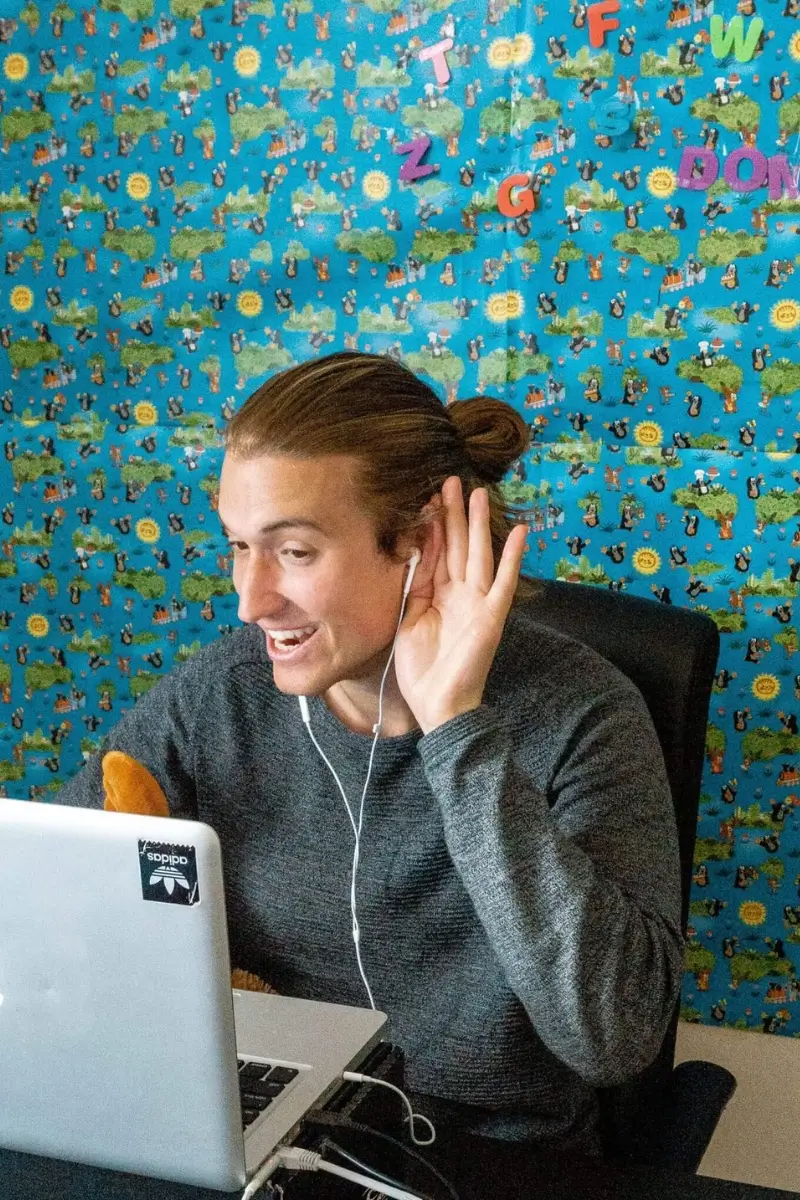 Dom teaching English online. He's sitting in front of his laptop listening to his student. He's got a postered background.