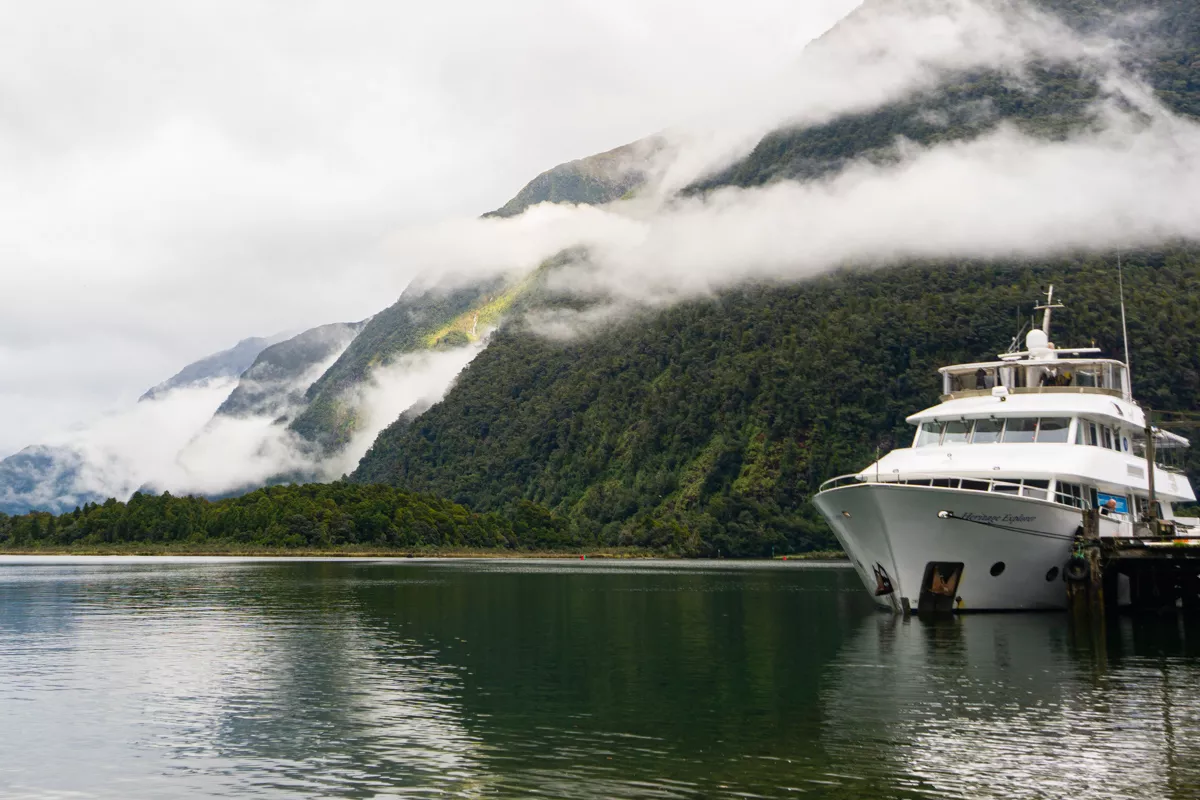 A white expedition yacht named “Heritage Explorer” is tied to a wooden jetty on Milford Sound’s glass-calm water, while low ribbons of cloud snake across the steep, forest-clad fiord walls in the background. The scene feels hushed and misty, with the boat’s bow reflecting faintly in the emerald water.