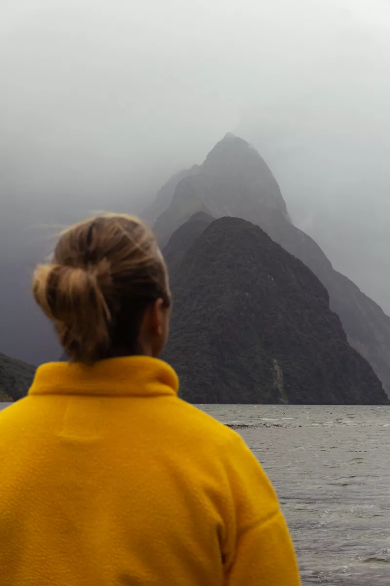 Viewed from behind, a person in a bright mustard-yellow fleece gazes across choppy water toward the dark, mist-shrouded spire of Mitre Peak rising into low cloud. The contrast between the vivid jacket and the brooding, foggy landscape draws the eye to the iconic mountain.