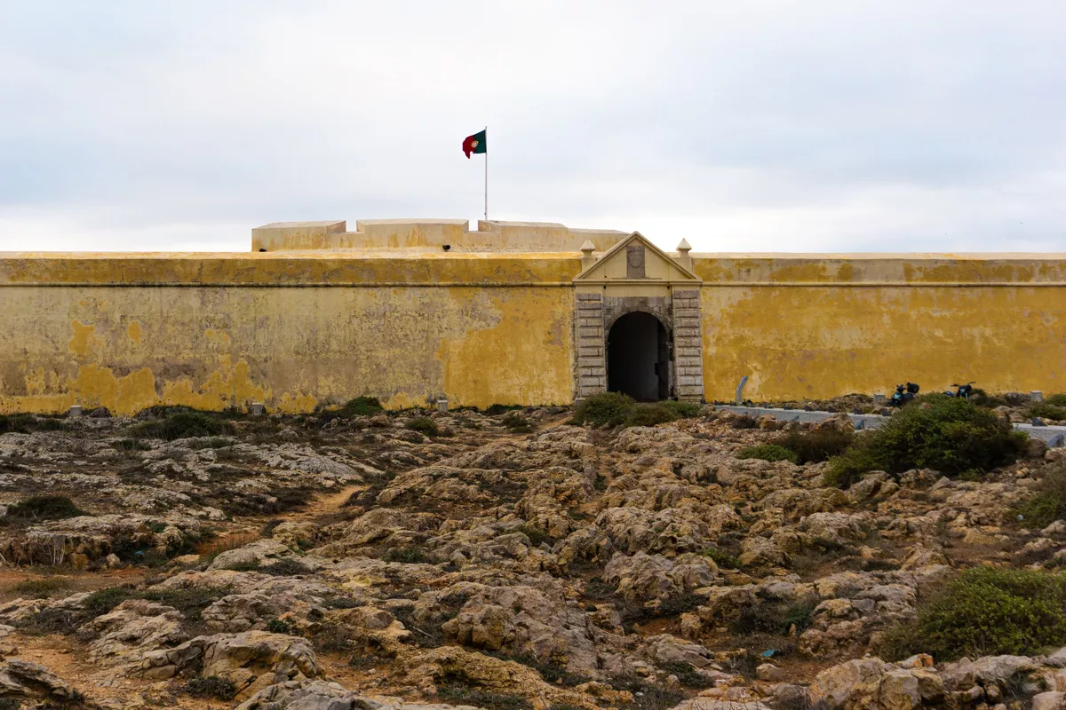 The entrance to a yellow Fortress in Sagres, Algarve on a cloudy day. There is a Portuguese flag on top of the fortress.