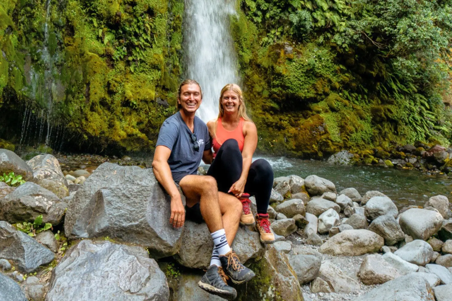 Smiling couple in hiking gear sitting on boulders with a lush, moss-covered waterfall cascading behind them on the Wilkies Pools & Dawson Falls Loop Trail in New Zealand.