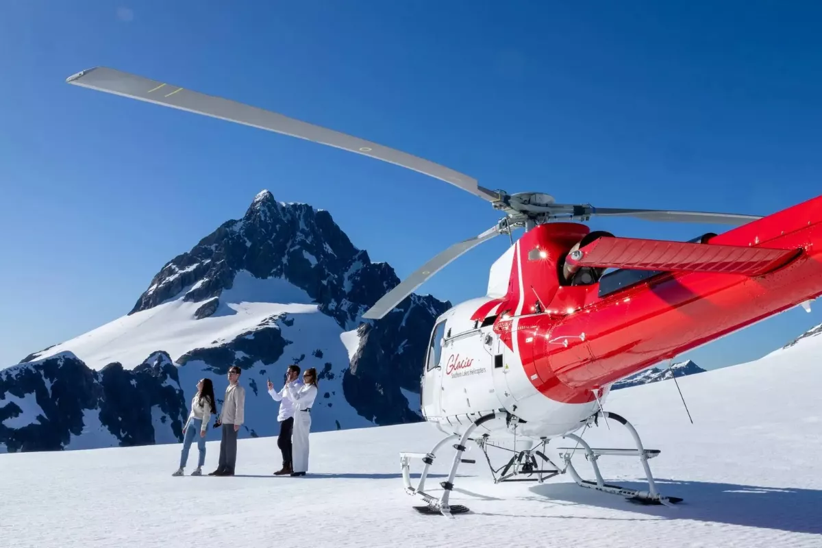 A bright red and white Glacier Southern Lakes Helicopters chopper on a snowy mountaintop under clear blue skies, with a dramatic snow-covered peak rising in the background. Four people stand nearby on the glacier, enjoying the view and taking photos.