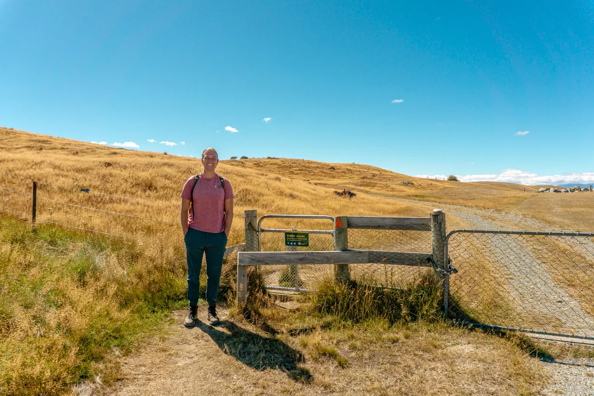 A smiling hiker stands at the Pukaki Kettle Hole Track trailhead next to a wooden fence and a metal gate marked “Public Access,” surrounded by golden grass under a bright blue sky. The trail begins behind the gate, winding through gently rolling hills.