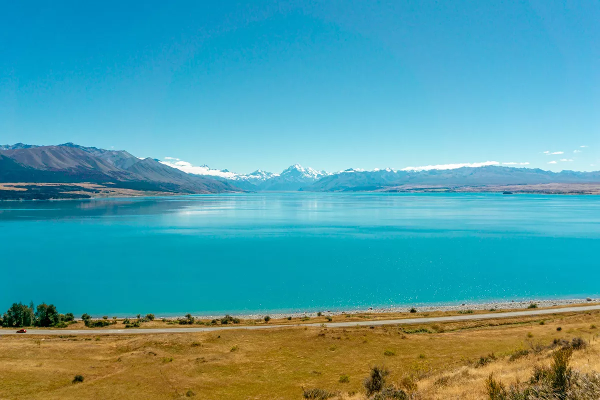A wide-angle view of Lake Pukaki with the Highway 8 road running along the shoreline and Mount Cook rising majestically in the distance. The scene is framed by golden grasslands and rugged mountain ranges beneath a clear blue sky.
