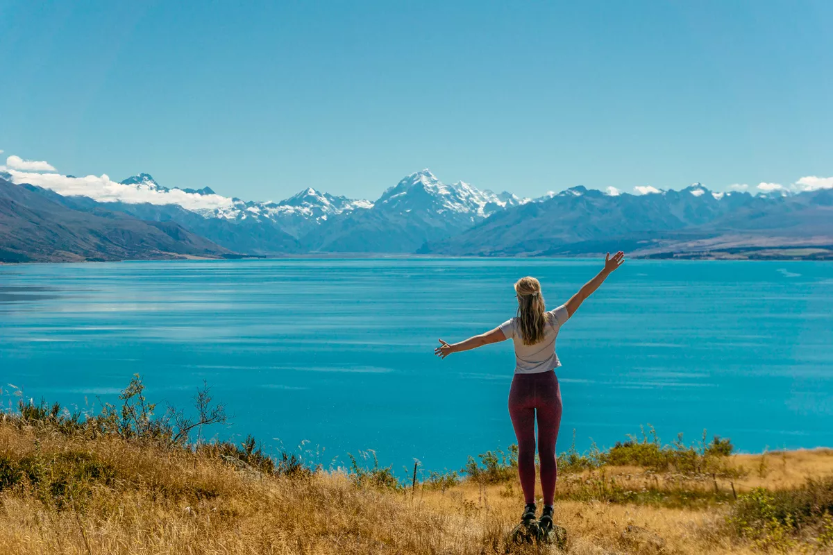 A woman stands on a grassy hill with arms outstretched, facing the vibrant turquoise waters of Lake Pukaki and the snow-capped peak of Mount Cook in the distance under a clear blue sky.