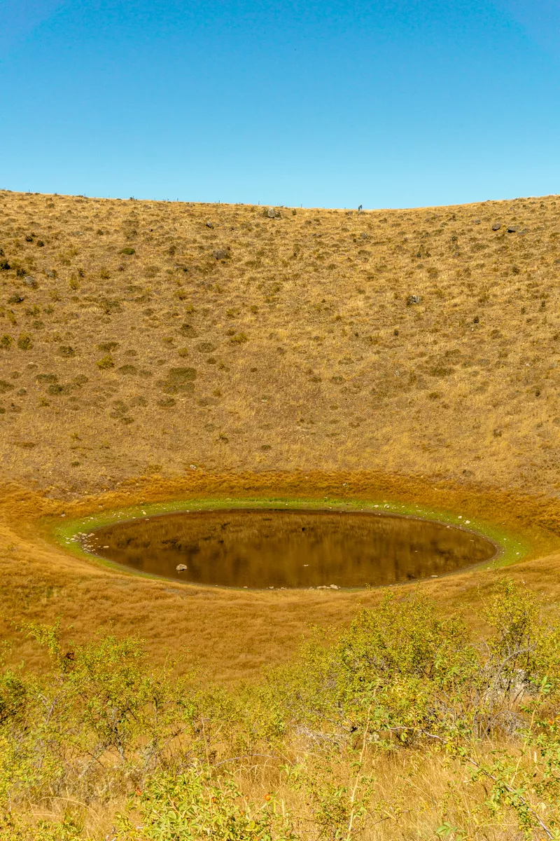 A circular body of water known as the Pukaki kettle hole sits carved into a dry, golden hillside, partially filled with still, dark water and ringed by a subtle band of green vegetation.