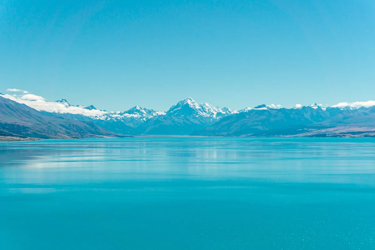 A breathtaking panoramic view of Lake Pukaki’s vivid turquoise waters stretches toward the snow-draped peak of Aoarki Mount Cook, surrounded by the rugged Southern Alps under a clear blue sky.