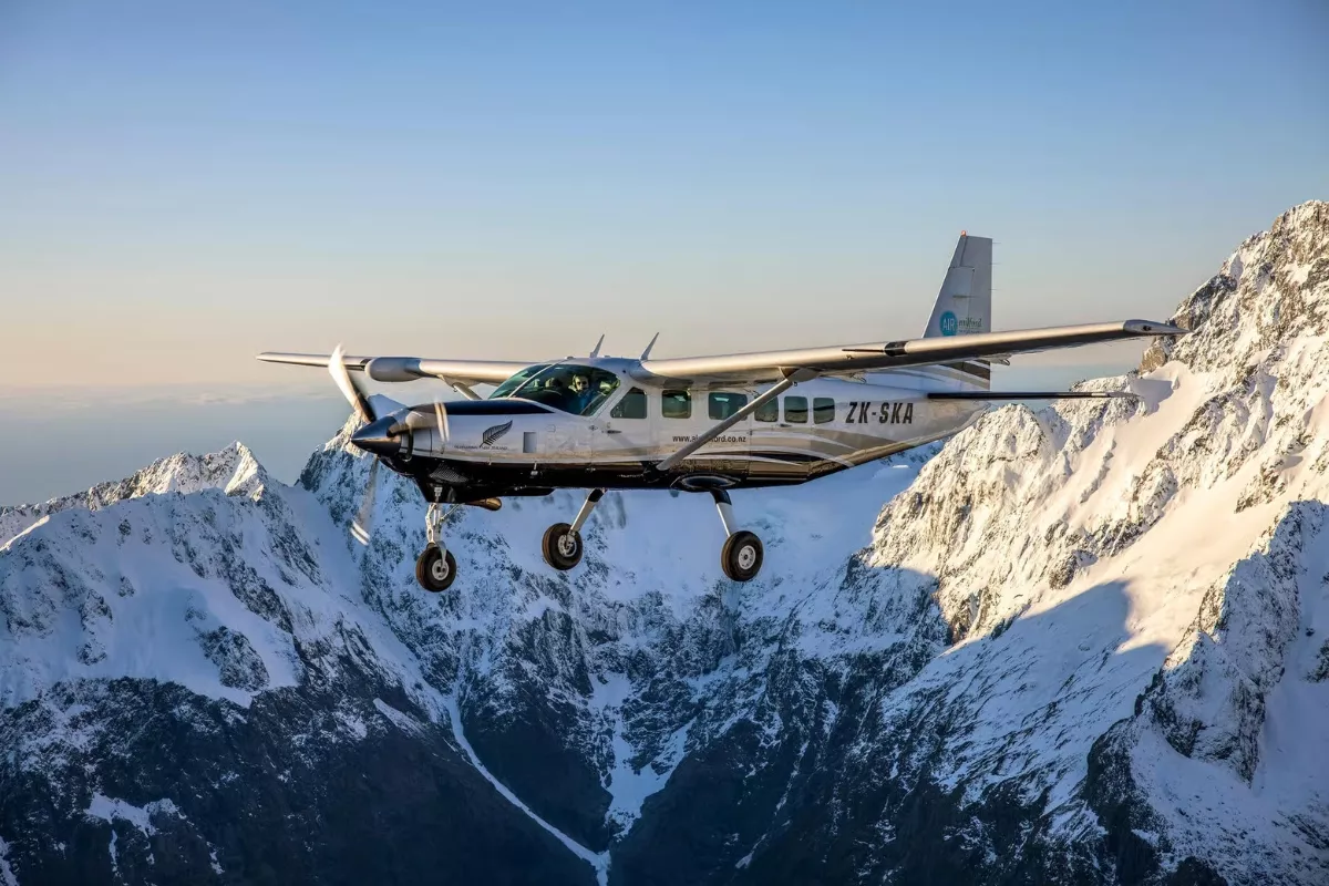 A small Air Milford aircraft with the flying past rugged, snow-covered peaks under a soft golden sky.