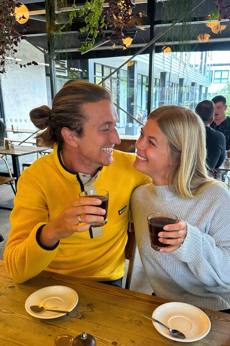 A smiling drinking coffee while sitting close together at a cozy café. The man wears a bright yellow fleece, and the woman wears a light blue sweater, both looking at each other with joy.