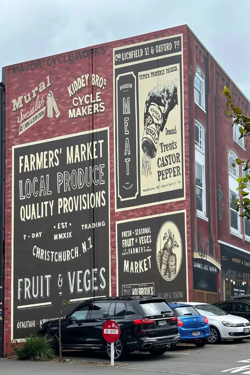 A large vintage-style mural on a brick building in Christchurch, New Zealand, advertising a local farmers' market with signs for fruit and vegetables, meat, and castor pepper. Several parked cars and a “No Entry” sign are visible in front of the mural.