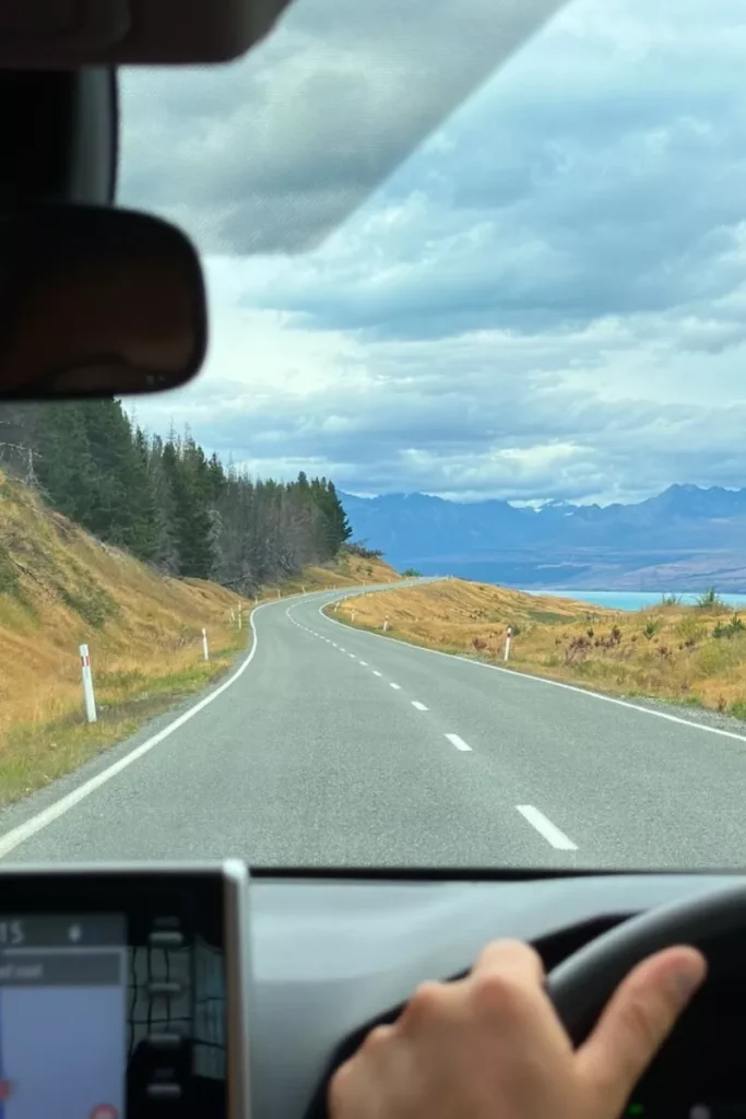 View from inside a car driving along the road to Aoraki/Mount Cook, curving through golden grasslands and pine trees with distant views of snow-capped mountains and a turquoise lake under a cloudy sky. A hand on the steering wheel and part of a GPS screen are visible, capturing the feeling of a scenic New Zealand road trip.