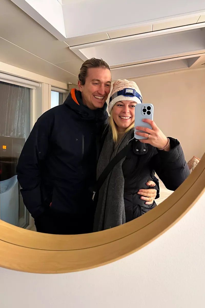 A smiling couple takes a mirror selfie indoors, both dressed in winter coats. The woman wears a white knit hat and holds a phone with a light blue case while the man has his arm around her waist.