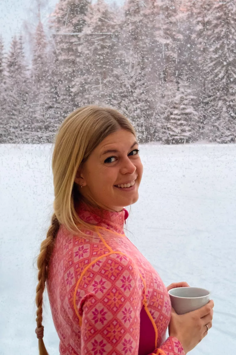 A woman in a bright pink Nordic-patterned base layer stands near a frosted window with snowy trees in the background. She smiles over her shoulder while holding a white mug.