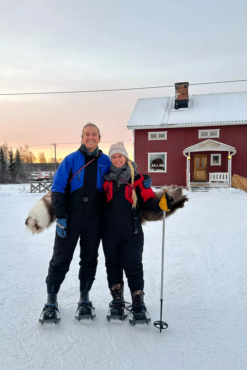 A smiling couple dressed in winter gear stands side by side on snowshoes in front of a red wooden cabin with a snow-covered roof. They each hold a rolled-up reindeer hide.