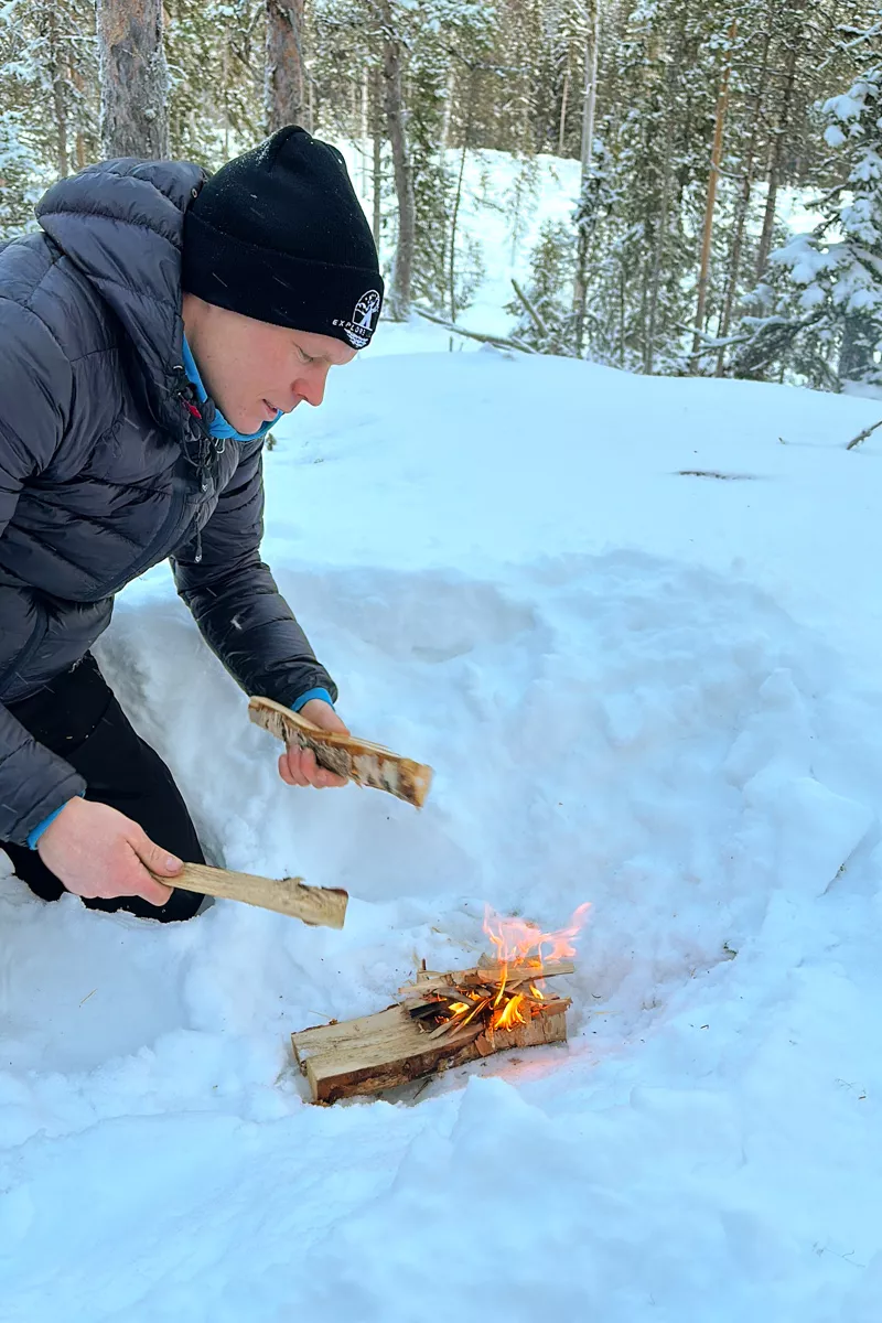A person in a black beanie and puffer jacket crouches in the snow while building a small fire with kindling.