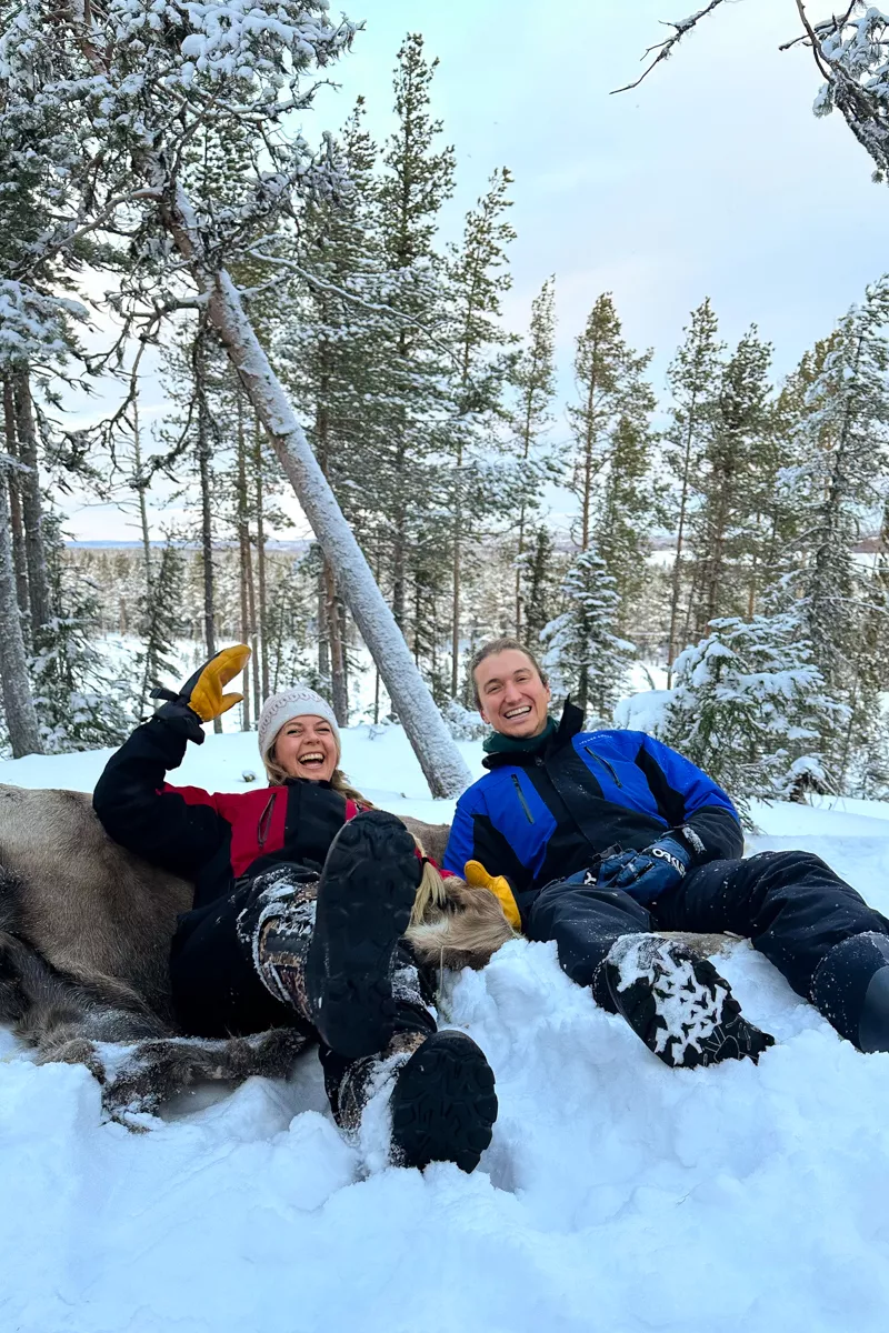 A couple lies back laughing in the snow on top of a reindeer hide, surrounded by snow-covered trees.