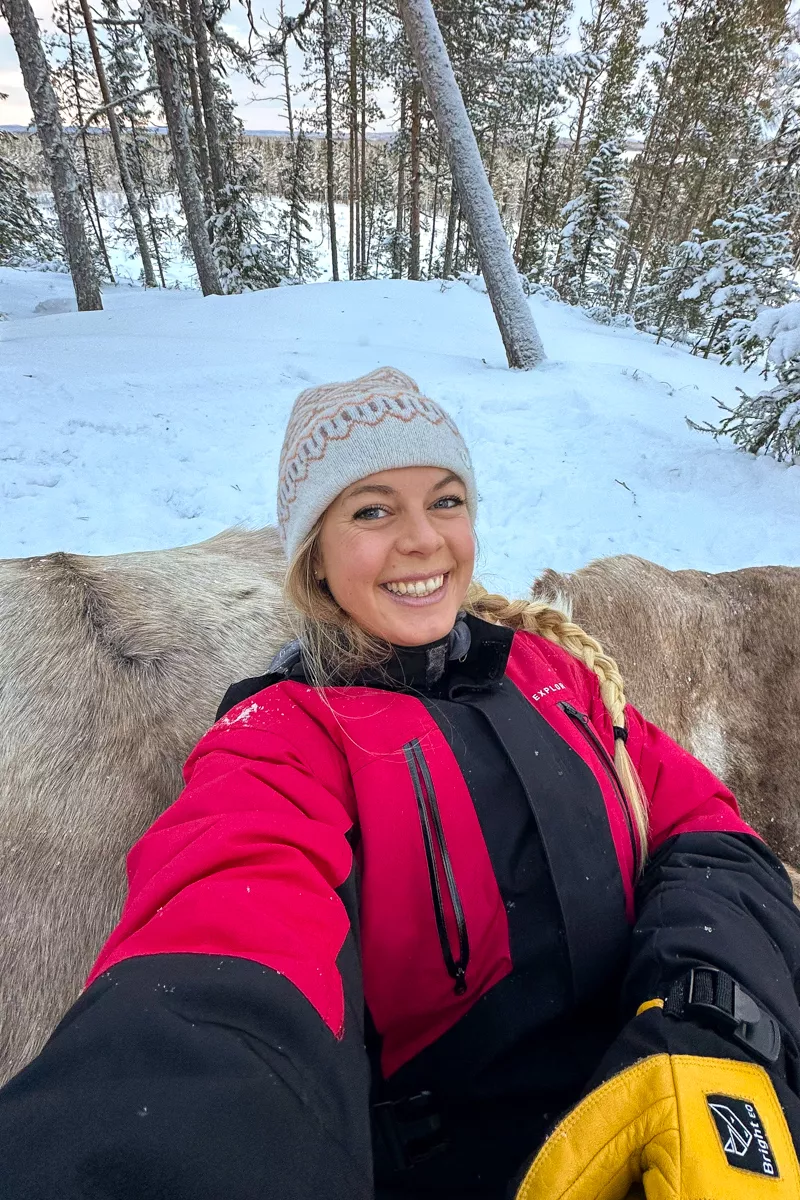 A woman in a red and black snowsuit smiles brightly while taking a selfie outdoors, sitting on a reindeer hide in a snowy forest.