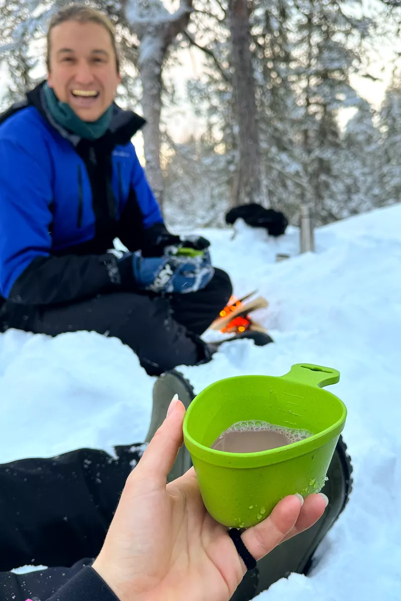 A close-up of a green camping cup filled with hot chocolate is held in the foreground while a man smiles in the snowy background near a small fire.