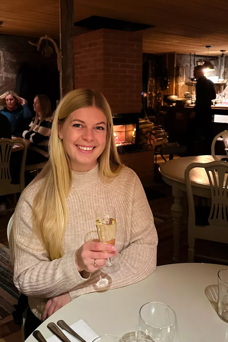 A woman with long blond hair smiles while holding a glass of white wine at a cozy, warmly lit restaurant.