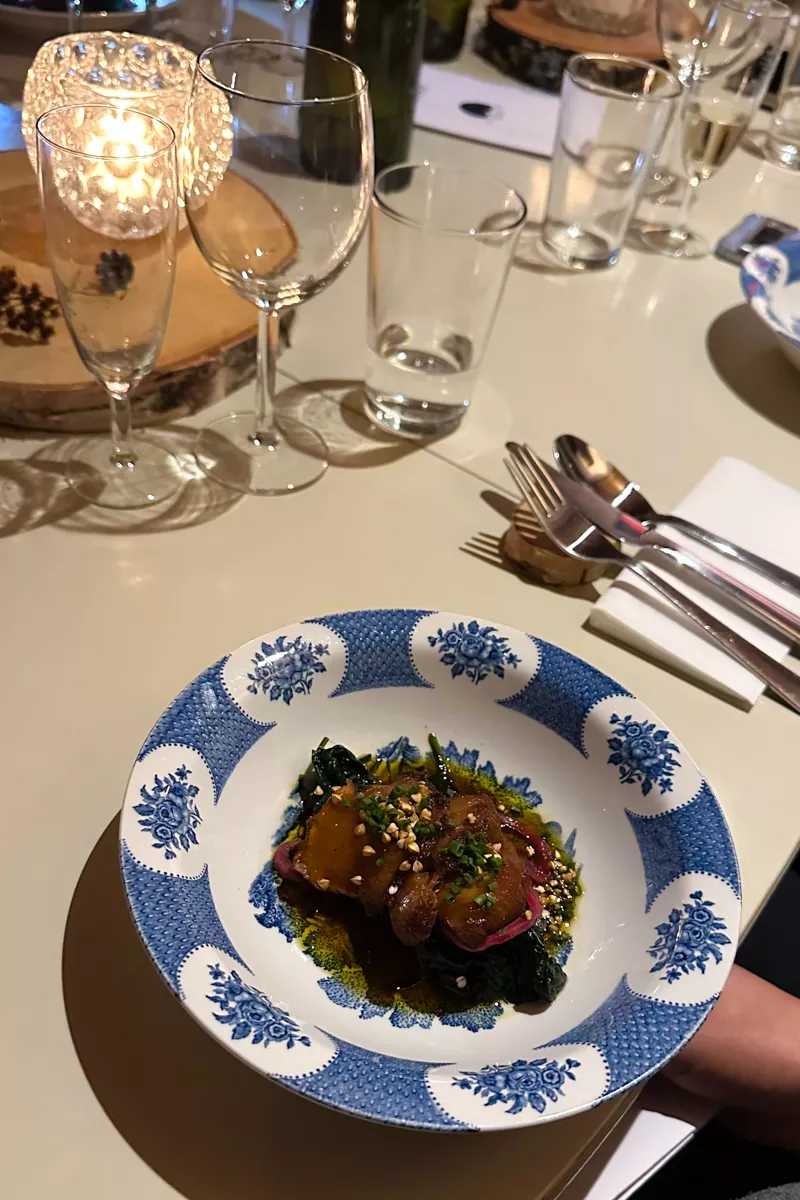 A beautifully plated dish featuring roasted vegetables and greens is served in a blue and white patterned plate. The table is set with wine glasses, cutlery, and a glowing candle centerpiece.