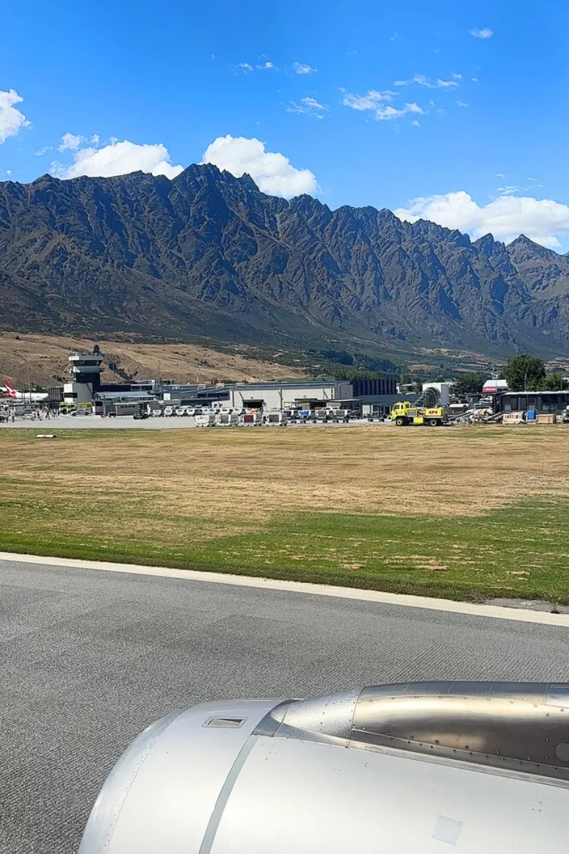 View from an airplane window of Queenstown Airport, with the rugged Remarkables mountain range in the background under a bright blue sky. The airport buildings and a yellow airport vehicle are visible near the runway.