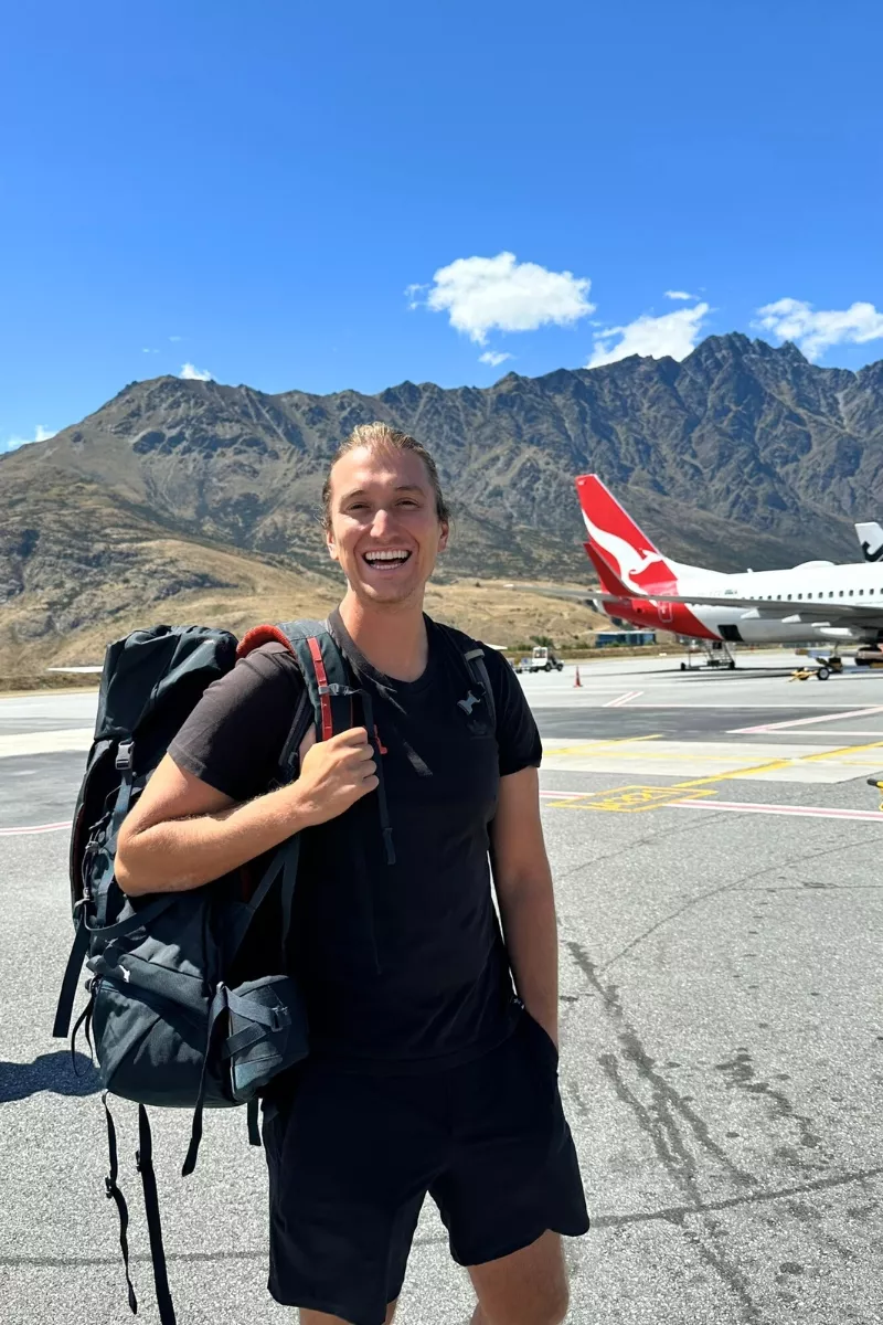 Smiling traveller standing on the tarmac at Queenstown Airport with a large backpack, wearing a black outfit, with a Qantas airplane and the Remarkables mountain range behind him under a sunny sky.