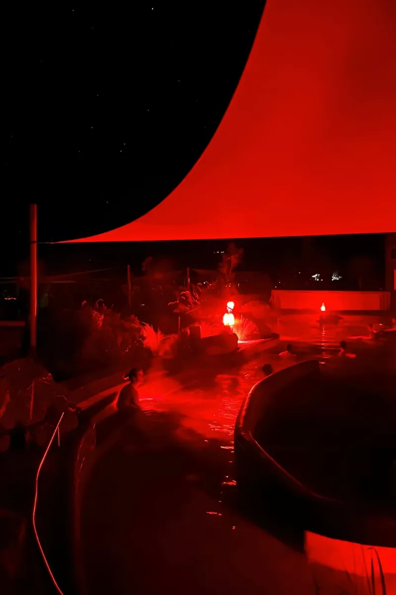 An outdoor hot spring at night illuminated by red lighting, with people relaxing in the steaming water under a triangular red canopy. The night sky with faint stars is visible above the setup, creating a surreal, otherworldly ambiance.