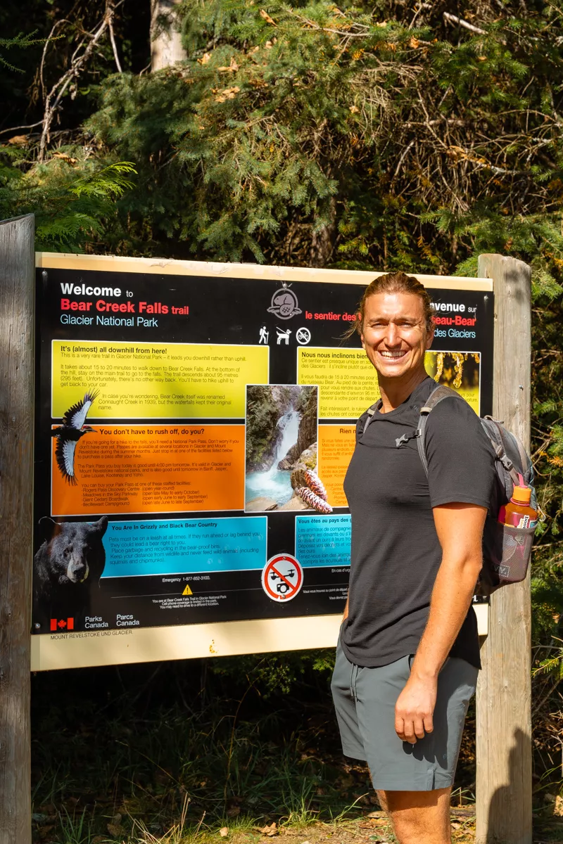 A smiling hiker stands in front of a colourful trailhead sign for Bear Creek Falls Trail in Glacier National Park. The sign provides safety tips, trail information, and a waterfall photo.