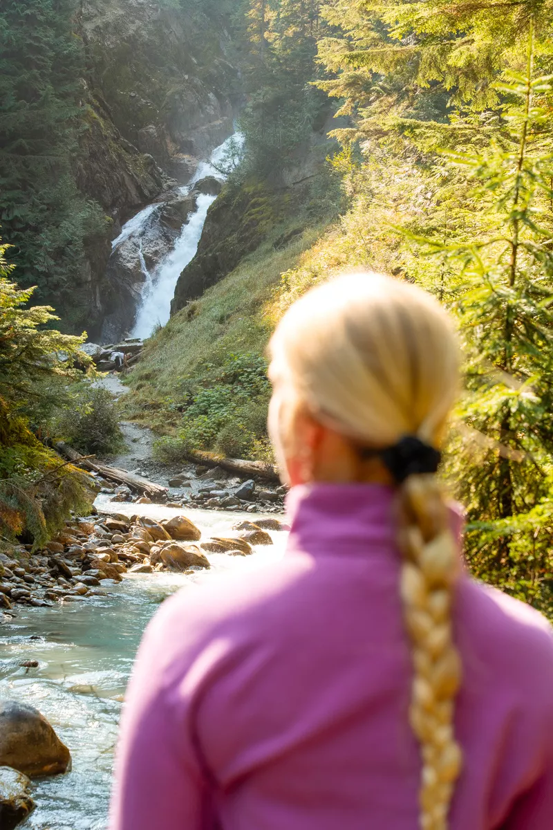 A woman in a pink sweater with a long blonde braid looks toward a distant waterfall framed by evergreens and a rocky stream. The shot is taken from behind her, capturing her perspective as she admires the natural landscape.